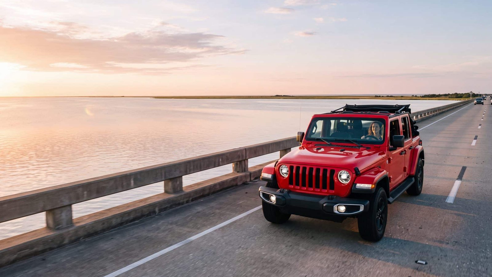 A red Jeep Wrangler JL Unlimited crossing a long low concrete highway bridge over a wide flat sound at golden hour, seen from the shoulder — Outer Banks, NC