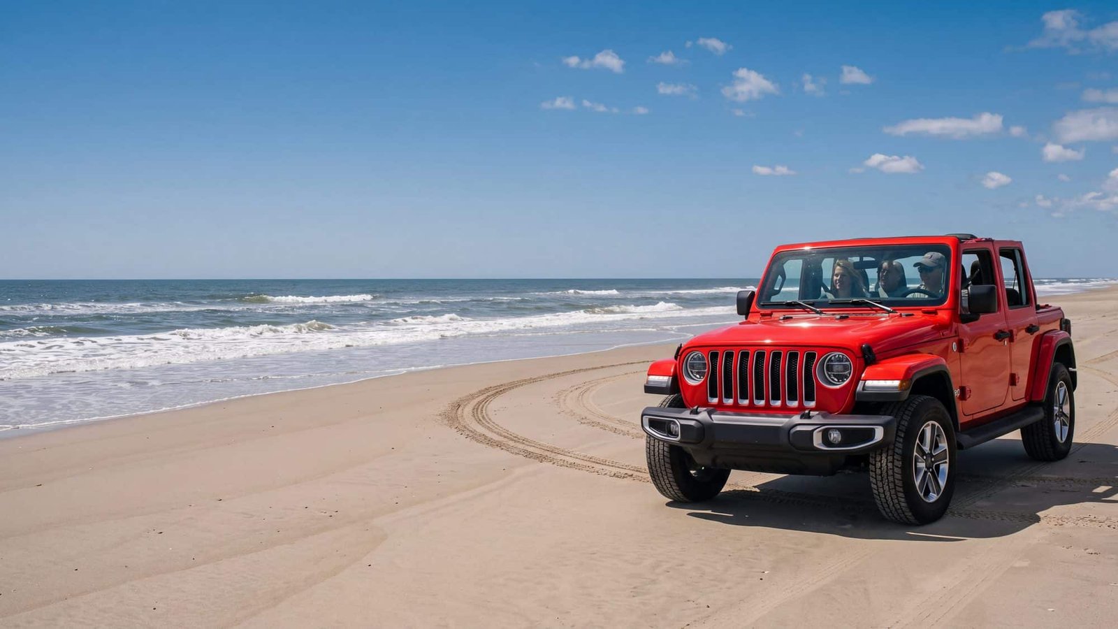 A red Jeep Wrangler JL Unlimited driving on the wide hard-packed sand at low tide with a couple inside seated and belted, tire tracks curving behind it — Outer Banks, NC