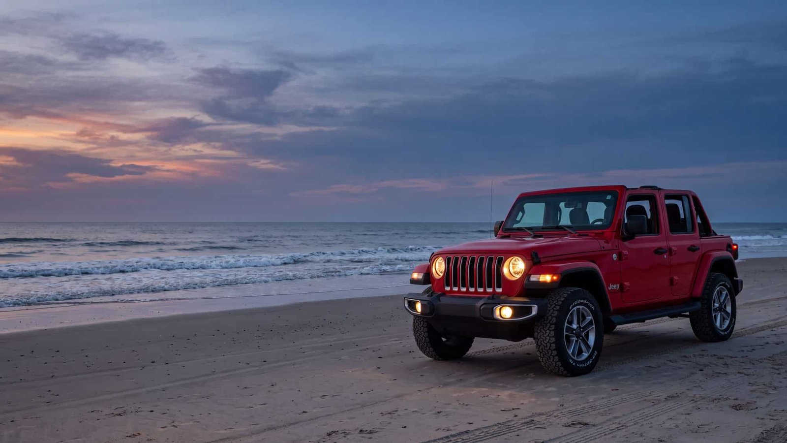 A red Jeep Wrangler JL Unlimited parked on hard sand facing the surf at sunset, empty — Outer Banks, NC